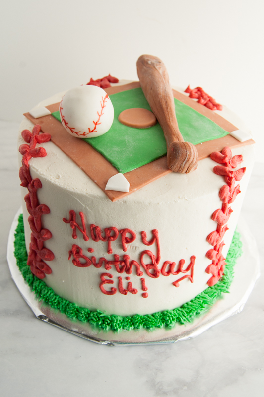 Round cake frosting with white icing and red piping made to look like baseball laces.  On the top of the cake is a fondant baseball diamond and a fondant baseball bat and ball