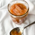 Jar of homemade applesauce on a white background with a spoon in front of the jar