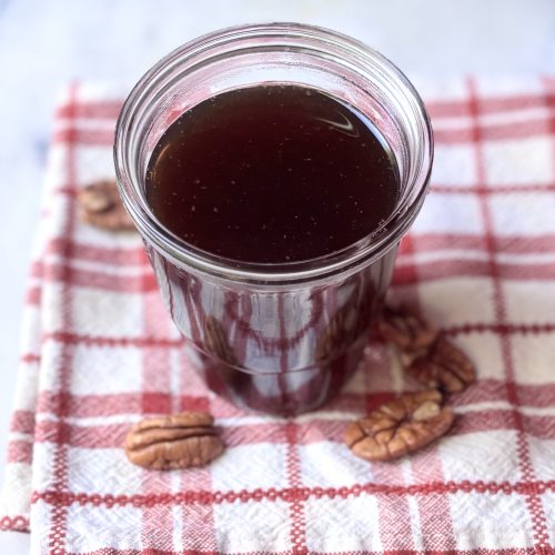 Jar of brown pecan brown sugar syrup on a red and white checked napkin surrounded by pecans