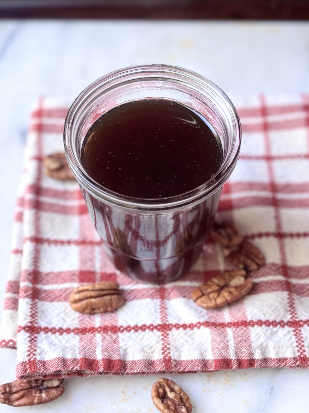 Jar of brown pecan brown sugar syrup on a red and white checked napkin surrounded by pecans