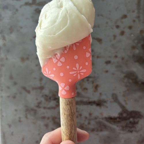 Cream cheese frosting on a pink spatula held up against a gray background