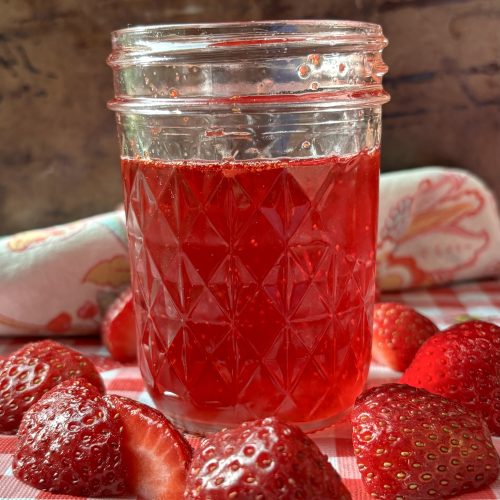 bright red strawberry syrup in a jar surrounded by fresh strawberries on a red and white checked table cloth