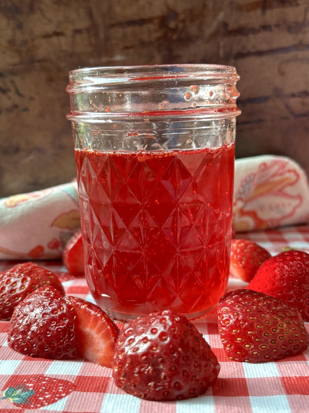 bright red strawberry syrup in a jar surrounded by fresh strawberries on a red and white checked table cloth